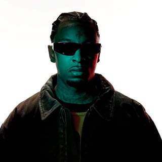 Portrait of a man wearing dark sunglasses and a denim jacket against a white background with teal and red lighting effects