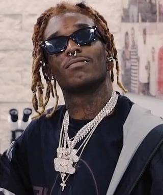 Man wearing dark sunglasses, diamond chain necklace, and black clothing with braided hair and hoop earrings