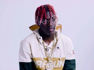 Portrait of a person with red braided hair wearing glasses, a white and gold jacket, and gold chain necklaces against a light background