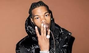Man with braided hair wearing a black shiny puffer jacket against a tan background, touching his face thoughtfully